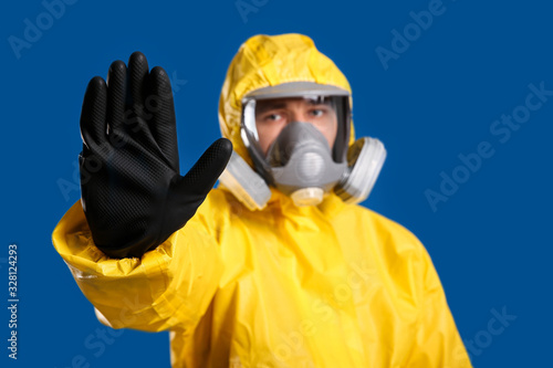 Man in chemical protective suit making stop gesture against blue background, focus on hand. Virus research