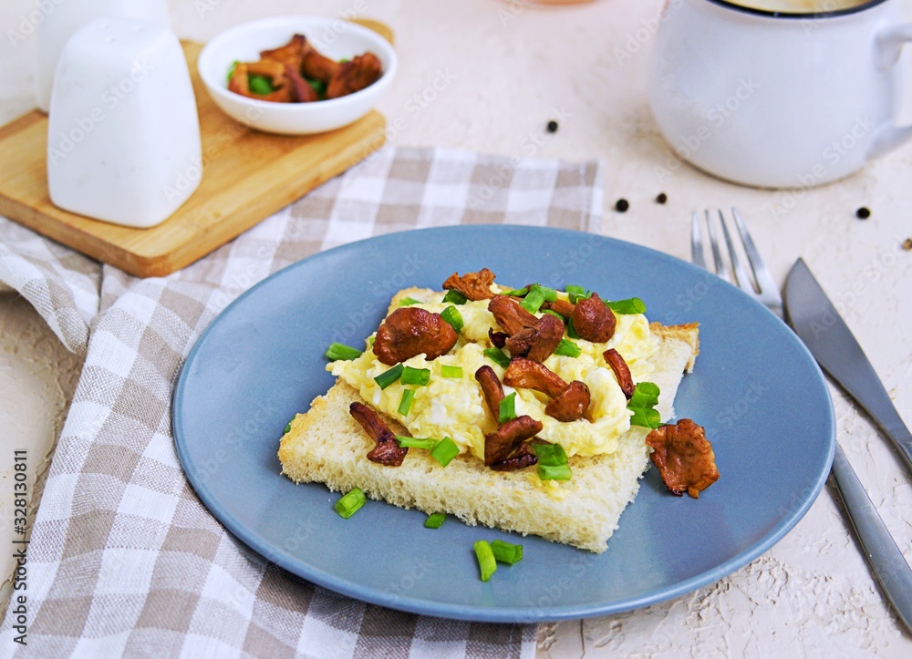 Tender scramble omelet on crispy white bread toast with fried chanterelles on a gray plate on a light concrete background. Tender scramble omelet on crispy white bread toast with fried chanterelles on a gray plate on a light concrete background.