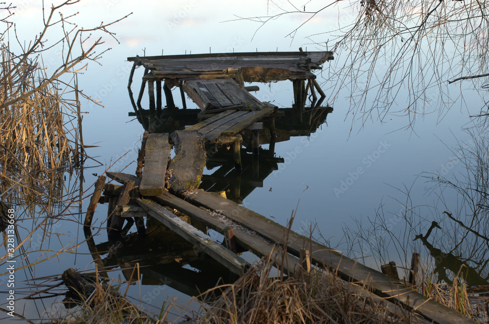 Spring landscape with Old destroyed wooden fishing dock on the river among the reeds. Abandoned pier or bridge for fishing with reflection in the water
