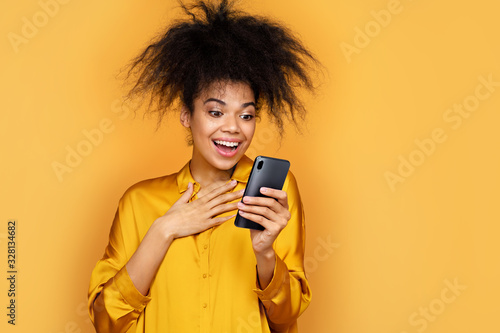 Young girl looks at the phone, happy to receive message. Photo of african american girl on yellow background