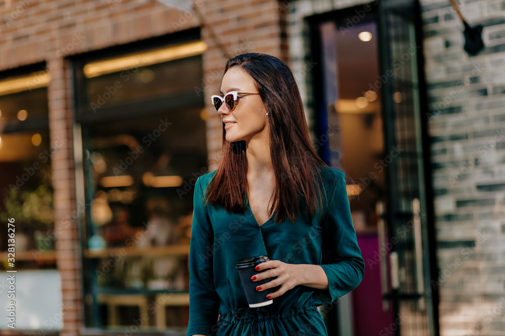 goodly girl in green dress and white glasses holding a cup of coffee smiling on the street. Well-dressed european woman spending time in city.