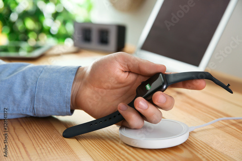 Man taking smartwatch from wireless charger at wooden table  closeup. Modern workplace accessory