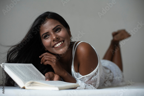 A young and attractive dark skinned Indian Bengali woman in lingerie reading book in a casual mood in white background. Indian lifestyle and  boudoir photography