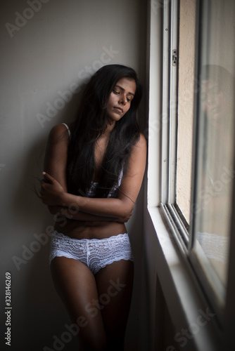Portrait of a young and attractive dark skinned Indian Bengali woman in lingerie posing in a casual mood in front of a window. Boudoir photography.