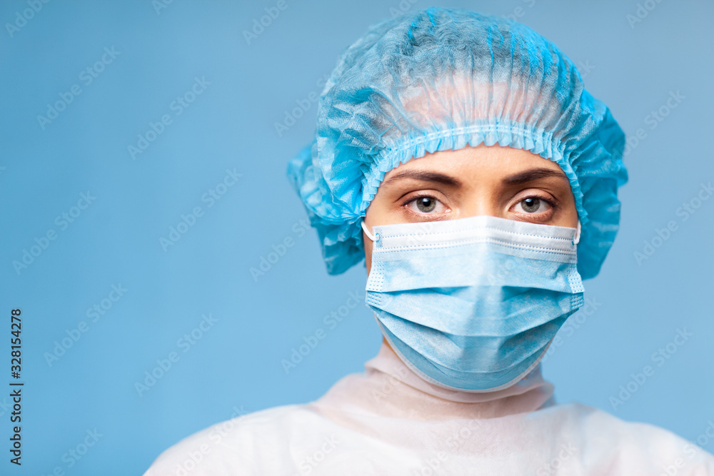 young female doctor in uniform, medical cap and face mask, close-up portrait. with a serious gaze looking at the camera on a blue background. copy space