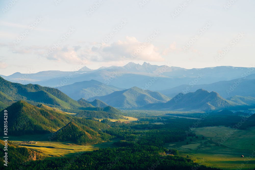 Wonderful mountain land with forest and small houses in sunlight. Scenic view to vast expanses of rolling hills and great mountain ridge on horizon. Immense distances of wavy relief in evening light.