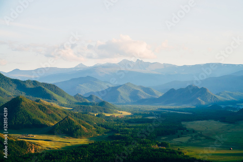 Wonderful mountain land with forest and small houses in sunlight. Scenic view to vast expanses of rolling hills and great mountain ridge on horizon. Immense distances of wavy relief in evening light.