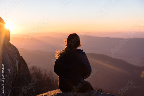 Amazing Landscape Photo With Silhouette of Woman on Hiking Adventure to Mountain Peak and Sitting on Rock Looking Out at Colorful Sunset Sky and Rolling Hills in Virginia with Sun Rays 