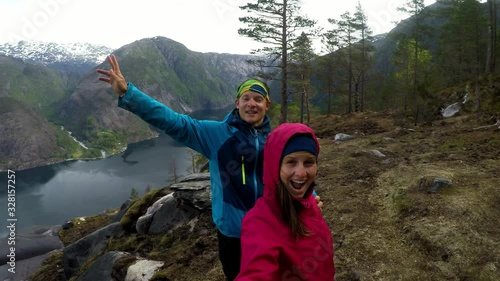 Couple standing next to Langfossen Waterfall and amazing fjord view on a cloudy day in Norway. The other side of the fjord is partially covered with snow. Navy blue water contrasted with green slopes photo