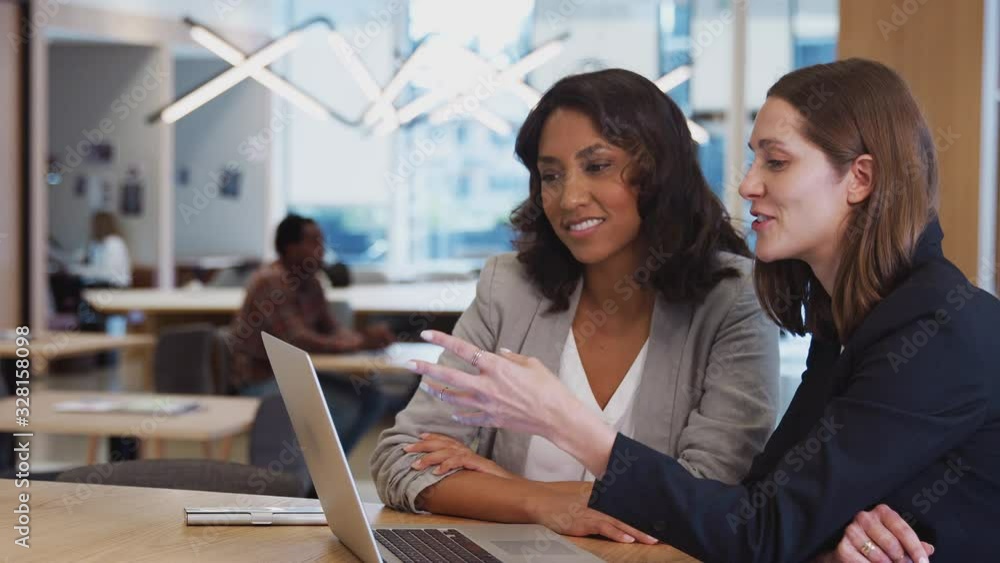 Two businesswomen with laptop at desk in open plan office collaborating on project together - shot in slow motion