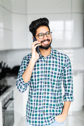 Happy indian young man in checkered shirt talking on cell phone on the kitchen