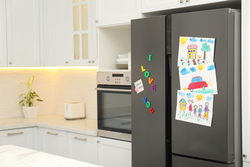 Modern refrigerator with child's drawings, note and magnets in kitchen. Space for text