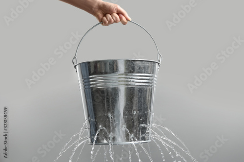 Woman holding leaky bucket with water on light grey background, closeup photo