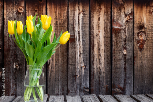Bunch of bright yellow spring tulips in a glass container on a rustic plank table.