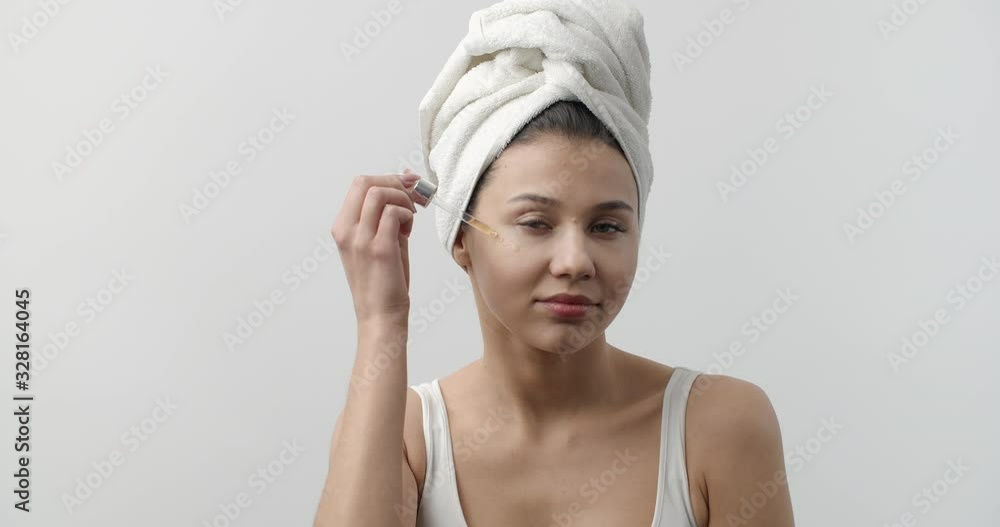 Portrait of pretty young lady in white bath towel applying serum on face with help of pipette. Sensual lady with perfect skin doing cosmetic procedure after shower. Portrait of pretty young lady in white bath towel applying serum on face with help of pipette. Sensual lady with perfect skin doing cosmetic procedure after shower.