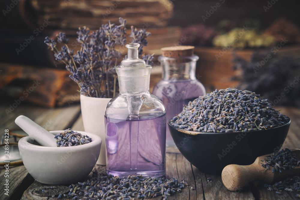 Bowl of dry lavender flowers, mortars, bottles of essential lavender oil or  infused water. Old books and medicinal herbs on background. Stock Photo