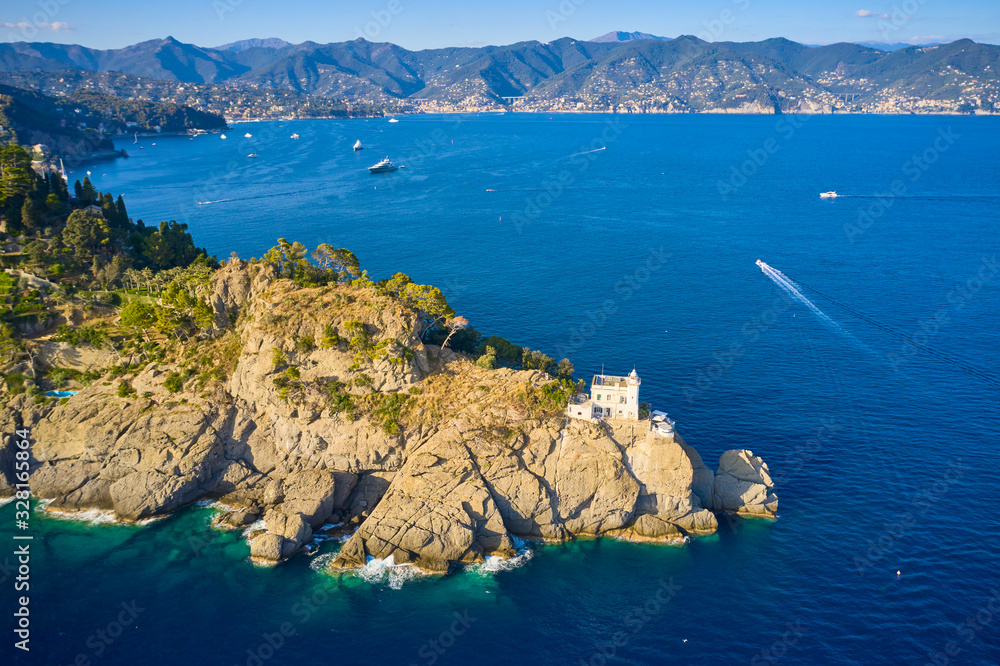 Top view on a lighthouse standing on the rocky hill near the sea with blue turquoise water in Portofino, Italy. Ligurian sea washes a coast with the big stones. Yachts are sailing near the coast.