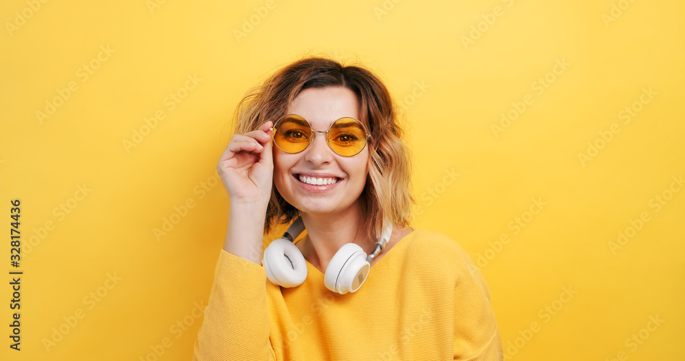 Portrait of happy young woman in white headphones correcting yellow glasses on her forehead coquettishly showing tongue camera smiling on isolated yellow background. Monotone. Emotions of people Portrait of happy young woman in white headphones correcting yellow glasses on her forehead coquettishly showing tongue camera smiling on isolated yellow background. Monotone. Emotions of people
