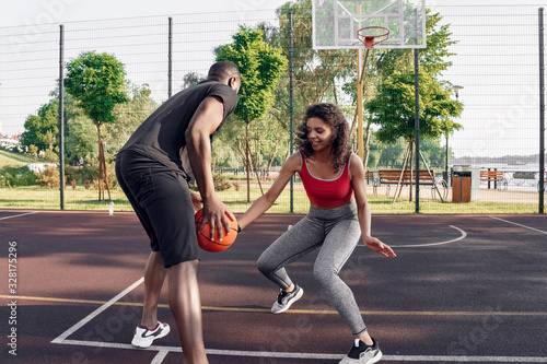 Outdoors Activity. African couple on basketball court guy dribbling while girl blocking smiling excited photo