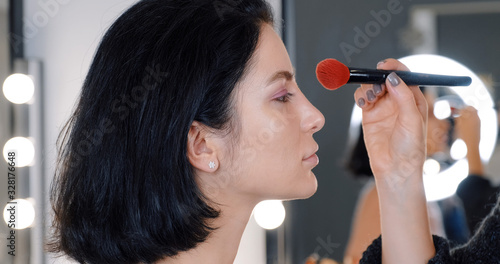 Portrait of brunette girl makeup artist on her face applying powder on her face with professional brush in makeup studio one background of bokeh light bulbs on mirror. Makeup and beauty. Lifestyle