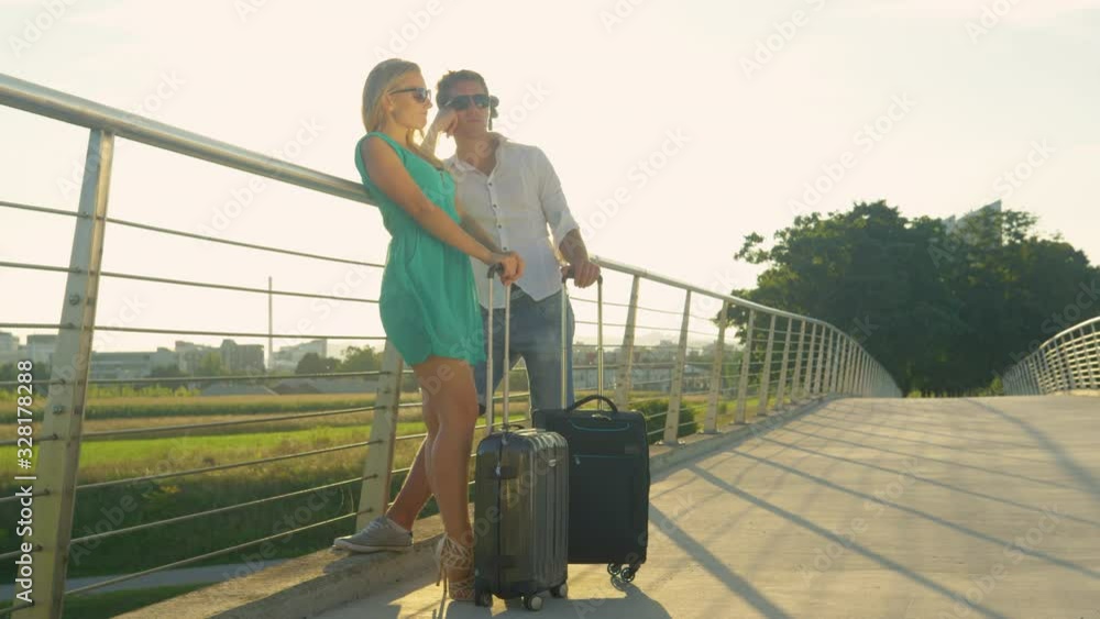 SLOW MOTION, COPY SPACE, LENS FLARE: Young newlyweds stand by side of the road waiting for the bus to take them to the airport. Traveler couple stand around waiting for the airport shuttle to arrive. SLOW MOTION, COPY SPACE, LENS FLARE: Young newlyweds stand by side of the road waiting for the bus to take them to the airport. Traveler couple stand around waiting for the airport shuttle to arrive.