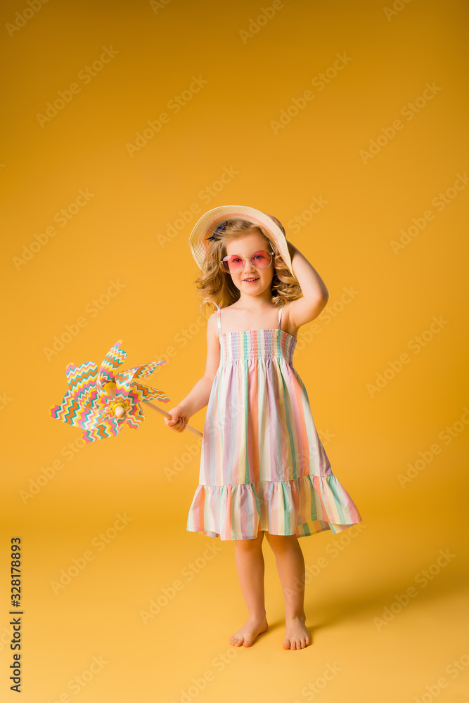 child girl blonde smiling in sunglasses and summer sundress holding a windmill stands on a yellow background. happy child. space for text child girl blonde smiling in sunglasses and summer sundress holding a windmill stands on a yellow background. happy child. space for text