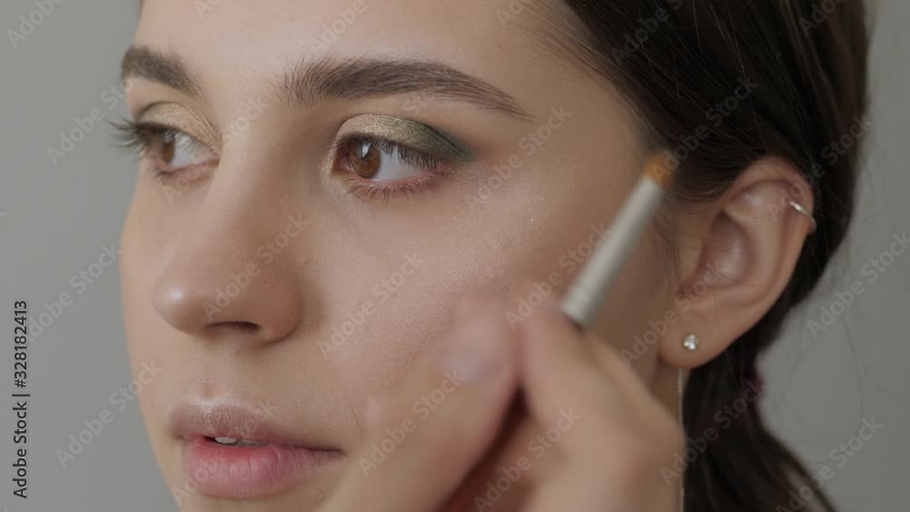 Big closeup slow motion shot of makeup master hand with a brush apply trendy professional make up golden smokey eyes in a beauty salon to a young adorable woman model with fresh skin indoors light Big closeup slow motion shot of makeup master hand with a brush apply trendy professional make up golden smokey eyes in a beauty salon to a young adorable woman model with fresh skin indoors light