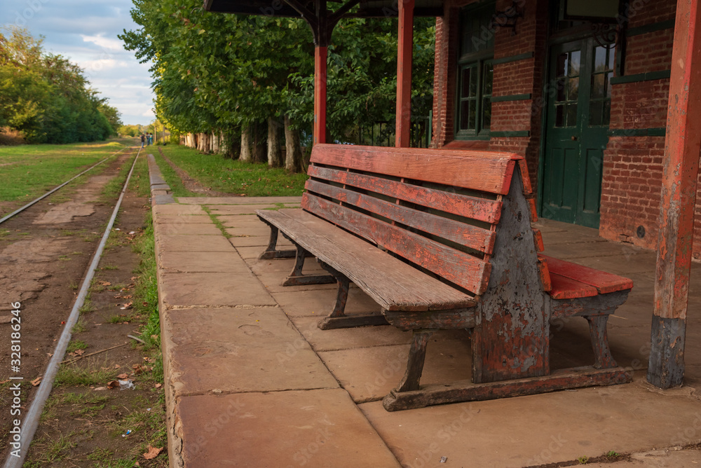 A wooden bench in an old abandoned train station with a view of the railroad with grass aroun it