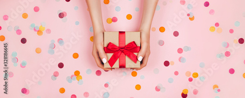Banner. Woman hands holding present box with red bow on pastel pink background with multicolored confetti. Flat lay style. photo