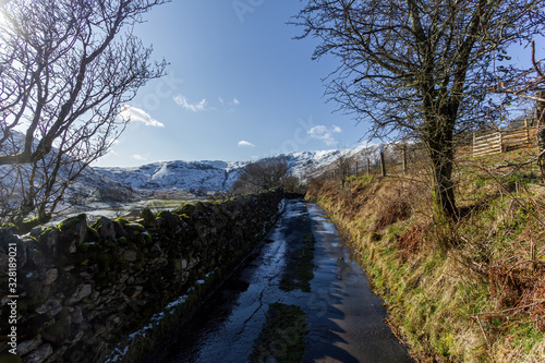 A scenic view of a wet rural road with snowy mountain range slope in the background under a majestic blue sky