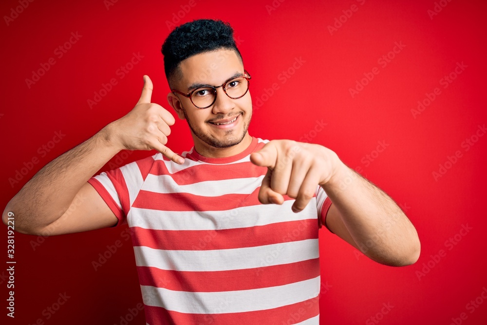 Young handsome man wearing casual striped t-shirt and glasses over isolated red background smiling doing talking on the telephone gesture and pointing to you. Call me. Young handsome man wearing casual striped t-shirt and glasses over isolated red background smiling doing talking on the telephone gesture and pointing to you. Call me.
