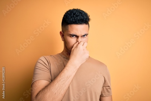 Young handsome man wearing casual t-shirt standing over isolated yellow background smelling something stinky and disgusting, intolerable smell, holding breath with fingers on nose. Bad smell © Krakenimages.com