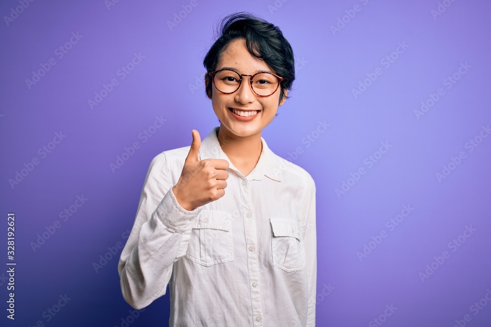 Young beautiful asian girl wearing casual shirt and glasses standing over purple background doing happy thumbs up gesture with hand. Approving expression looking at the camera showing success. Young beautiful asian girl wearing casual shirt and glasses standing over purple background doing happy thumbs up gesture with hand. Approving expression looking at the camera showing success.