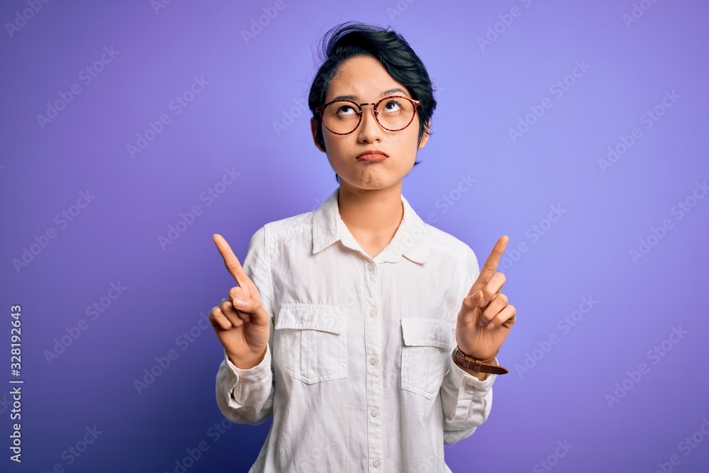 Young beautiful asian girl wearing casual shirt and glasses standing over purple background Pointing up looking sad and upset, indicating direction with fingers, unhappy and depressed. Young beautiful asian girl wearing casual shirt and glasses standing over purple background Pointing up looking sad and upset, indicating direction with fingers, unhappy and depressed.