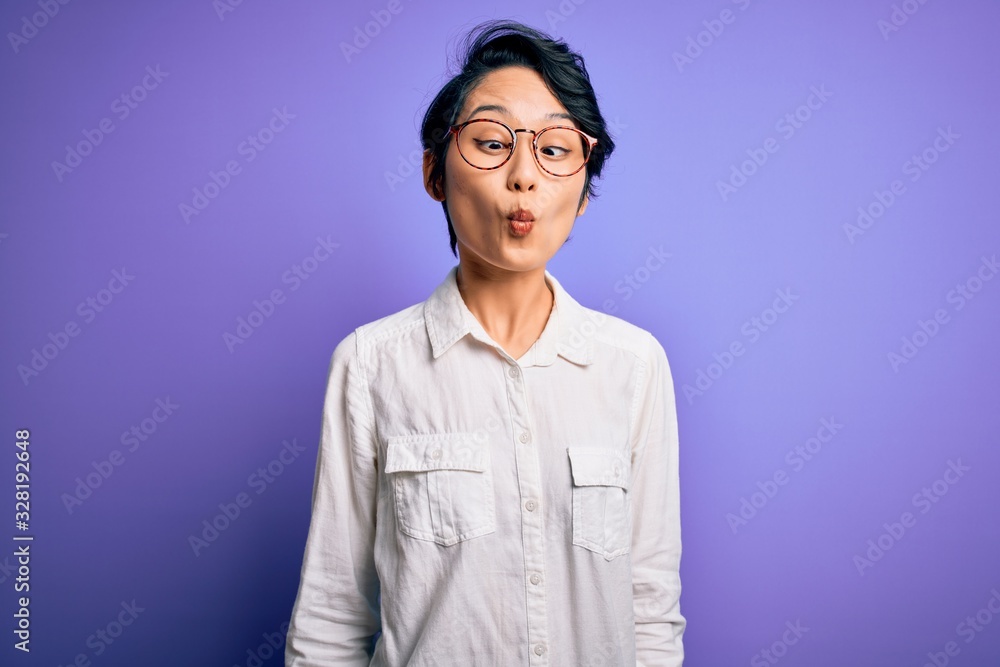 Young beautiful asian girl wearing casual shirt and glasses standing over purple background making fish face with lips, crazy and comical gesture. Funny expression. Young beautiful asian girl wearing casual shirt and glasses standing over purple background making fish face with lips, crazy and comical gesture. Funny expression.