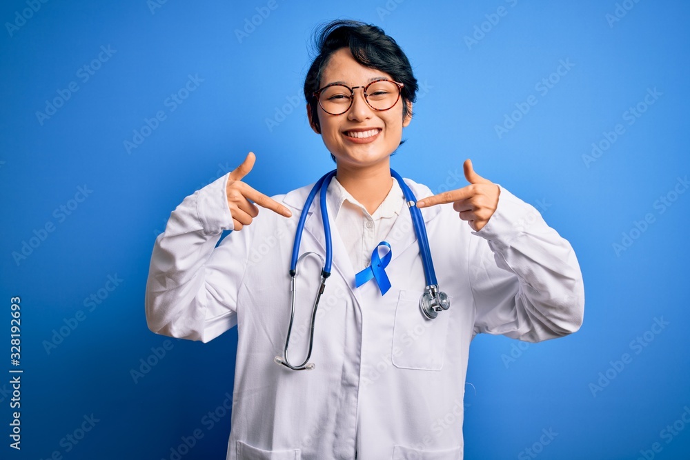 Young beautiful asian doctor girl wearing stethoscope and coat with blue cancer ribbon looking confident with smile on face, pointing oneself with fingers proud and happy. Young beautiful asian doctor girl wearing stethoscope and coat with blue cancer ribbon looking confident with smile on face, pointing oneself with fingers proud and happy.