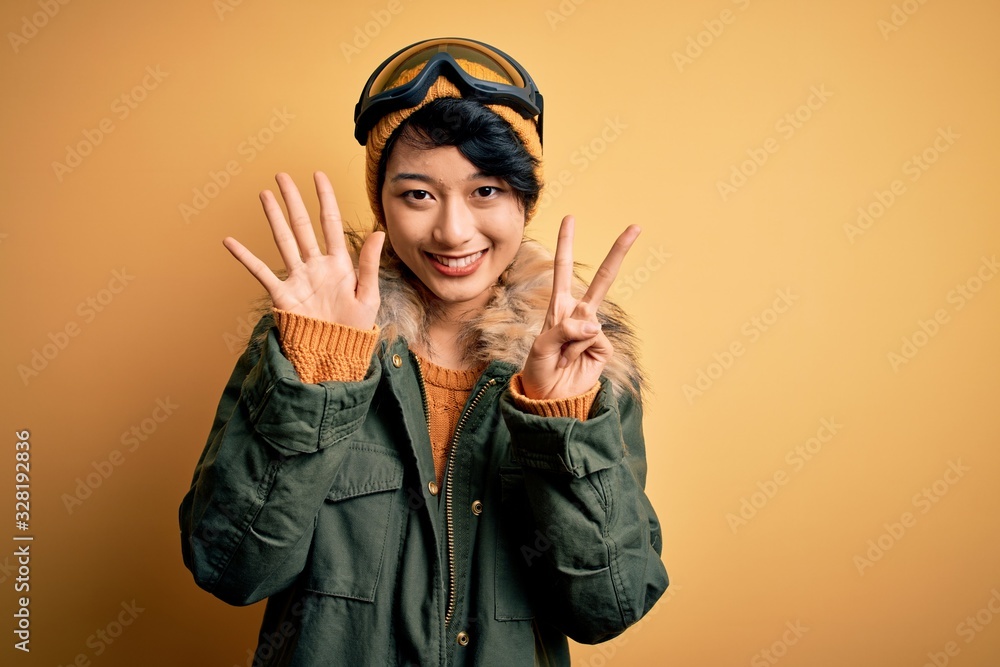 Beautiful asian skier girl wearing snow sportswear using ski goggles over yellow background showing and pointing up with fingers number seven while smiling confident and happy. Beautiful asian skier girl wearing snow sportswear using ski goggles over yellow background showing and pointing up with fingers number seven while smiling confident and happy.