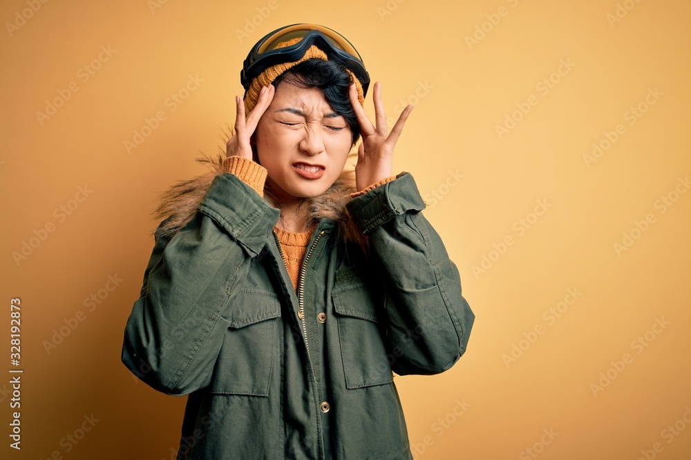 Beautiful asian skier girl wearing snow sportswear using ski goggles over yellow background with hand on head for pain in head because stress. Suffering migraine. Beautiful asian skier girl wearing snow sportswear using ski goggles over yellow background with hand on head for pain in head because stress. Suffering migraine.