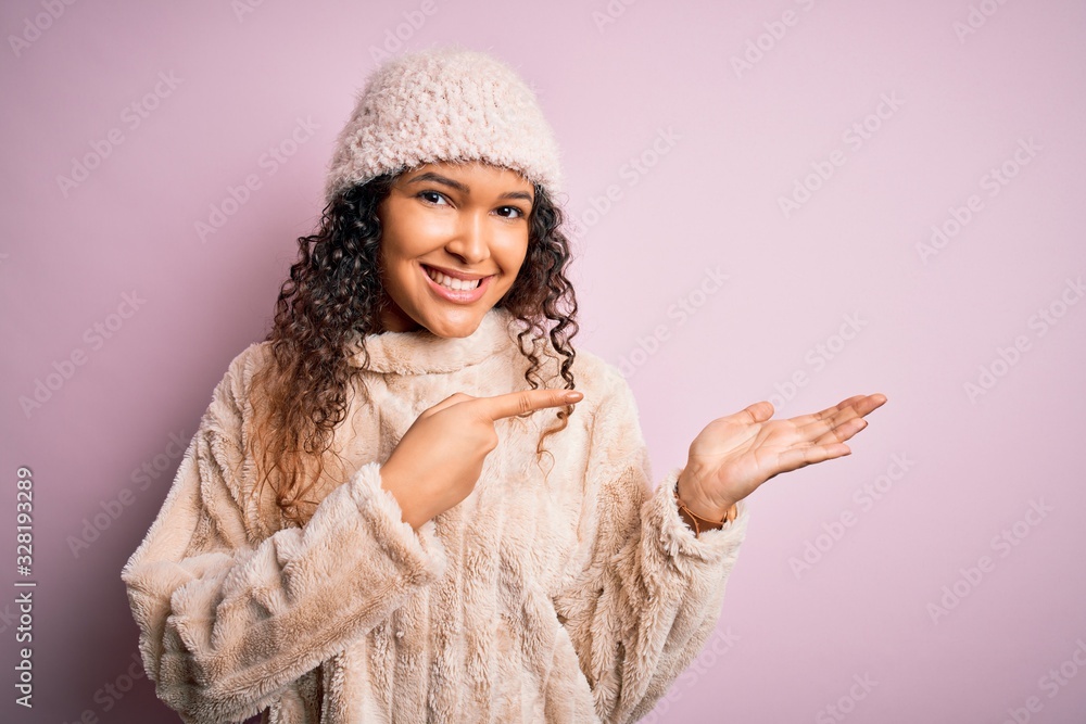 Beautiful woman with curly hair wearing casual sweater and wool cap over pink background amazed and smiling to the camera while presenting with hand and pointing with finger. Beautiful woman with curly hair wearing casual sweater and wool cap over pink background amazed and smiling to the camera while presenting with hand and pointing with finger.