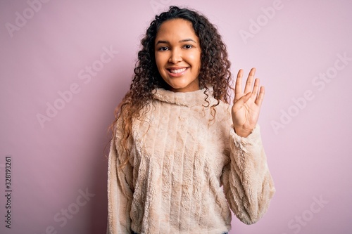 Young beautiful woman with curly hair wearing casual sweater standing over pink background showing and pointing up with fingers number four while smiling confident and happy.