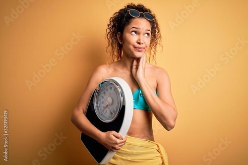 Young beautiful american slim woman on vacation wearing bikini holding weight machine serious face thinking about question  very confused idea