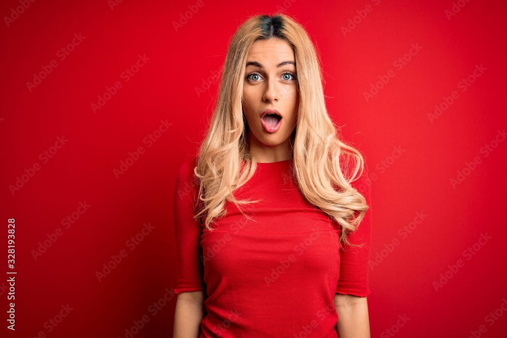 Young beautiful blonde woman wearing casual t-shirt standing over isolated red background afraid and shocked with surprise and amazed expression, fear and excited face. Young beautiful blonde woman wearing casual t-shirt standing over isolated red background afraid and shocked with surprise and amazed expression, fear and excited face.