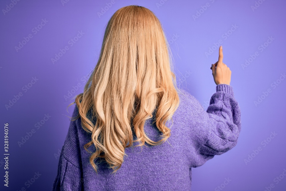 Young beautiful blonde woman wearing casual turtleneck sweater over purple background Posing backwards pointing ahead with finger hand Young beautiful blonde woman wearing casual turtleneck sweater over purple background Posing backwards pointing ahead with finger hand