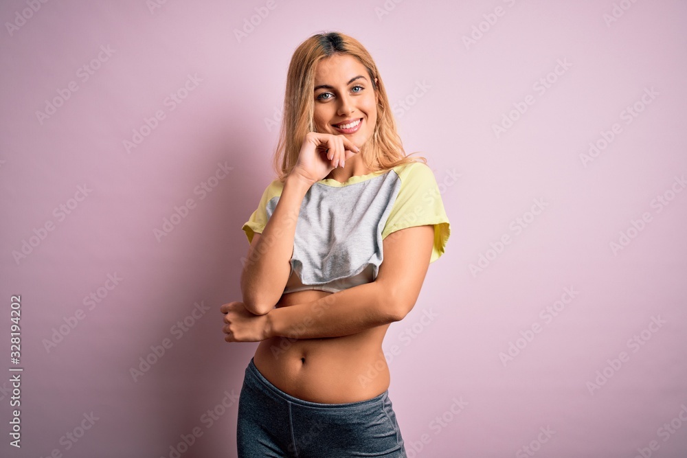 Young beautiful blonde sportswoman doing sport wearing sportswear over pink background looking confident at the camera with smile with crossed arms and hand raised on chin. Thinking positive. Young beautiful blonde sportswoman doing sport wearing sportswear over pink background looking confident at the camera with smile with crossed arms and hand raised on chin. Thinking positive.