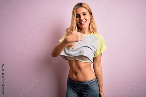 Young beautiful blonde sportswoman doing sport wearing sportswear over pink background doing happy thumbs up gesture with hand. Approving expression looking at the camera showing success.