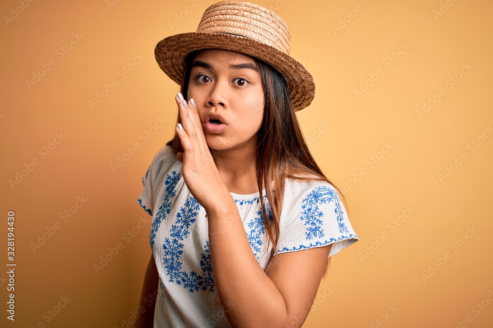 Young beautiful asian girl wearing casual t-shirt and hat standing over yellow background hand on mouth telling secret rumor, whispering malicious talk conversation Young beautiful asian girl wearing casual t-shirt and hat standing over yellow background hand on mouth telling secret rumor, whispering malicious talk conversation