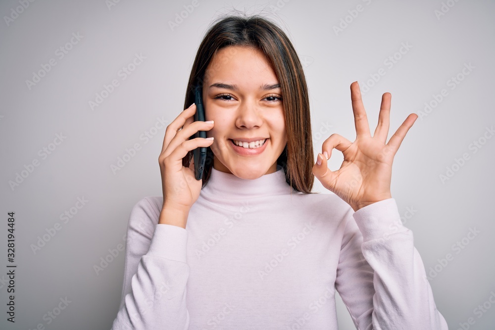 Beautiful brunette girl having conversation talking on the smartphone over white background doing ok sign with fingers, excellent symbol Beautiful brunette girl having conversation talking on the smartphone over white background doing ok sign with fingers, excellent symbol