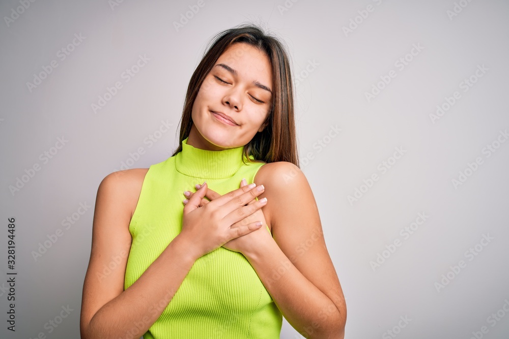 Young beautiful brunette girl wearing casual summer t-shirt over isolated white background smiling with hands on chest with closed eyes and grateful gesture on face. Health concept. Young beautiful brunette girl wearing casual summer t-shirt over isolated white background smiling with hands on chest with closed eyes and grateful gesture on face. Health concept.