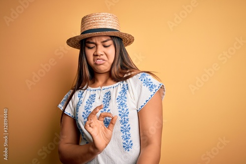 Young beautiful asian girl wearing casual t-shirt and hat standing over yellow background disgusted expression, displeased and fearful doing disgust face because aversion reaction.