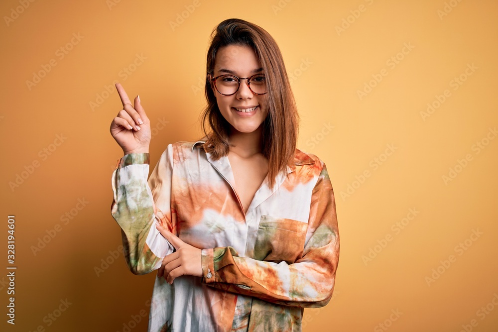 Young beautiful brunette girl wearing casual shirt and glasses over isolated yellow background with a big smile on face, pointing with hand and finger to the side looking at the camera. Young beautiful brunette girl wearing casual shirt and glasses over isolated yellow background with a big smile on face, pointing with hand and finger to the side looking at the camera.